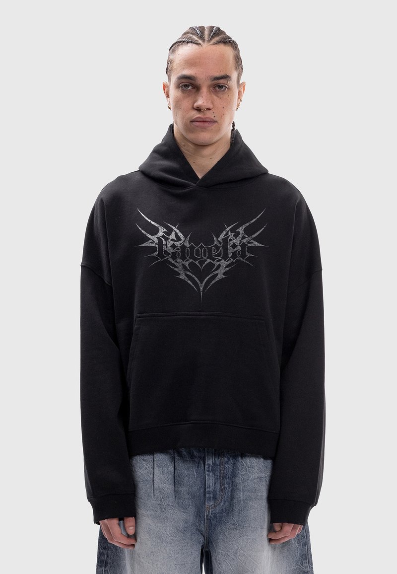 Personne aux cheveux tressés portant un sweat à capuche noir avec un motif gothique argenté et un jean bleu clair délavé, debout devant un fond gris uni.