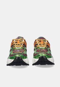 Vista frontal de zapatillas deportivas multicolores con acentos en verde, naranja, rojo y blanco, cordones beige y suelas negras sobre un fondo blanco.