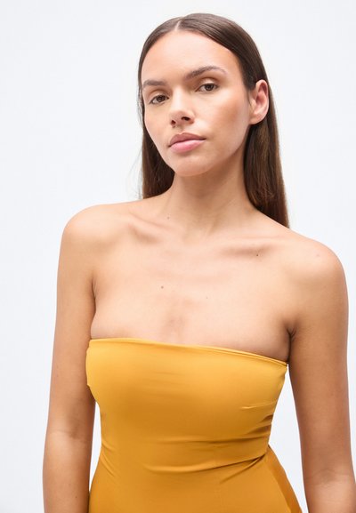 Κίτρινο φόρεμα χωρίς τιράντες, φτιαγμένο από απαλό ύφασμα, με εφαρμοστό σχέδιο που αγκαλιάζει τον κορμό, με ελάχιστες ραφές και ίσια γραμμή στο λαιμό.