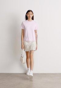 Lyserød bomulds t-shirt med hvid tekst, parret med en lys beige nederdel og hvide sneakers. En lille beige håndtaske holdes i venstre hånd.