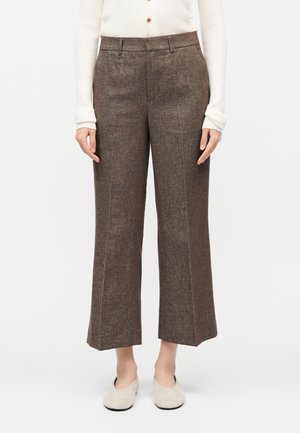 Femme portant un pantalon en tweed marron taille haute à jambes larges, un haut blanc à manches longues boutonné, et des chaussures beige slip-on, debout sur fond blanc.
