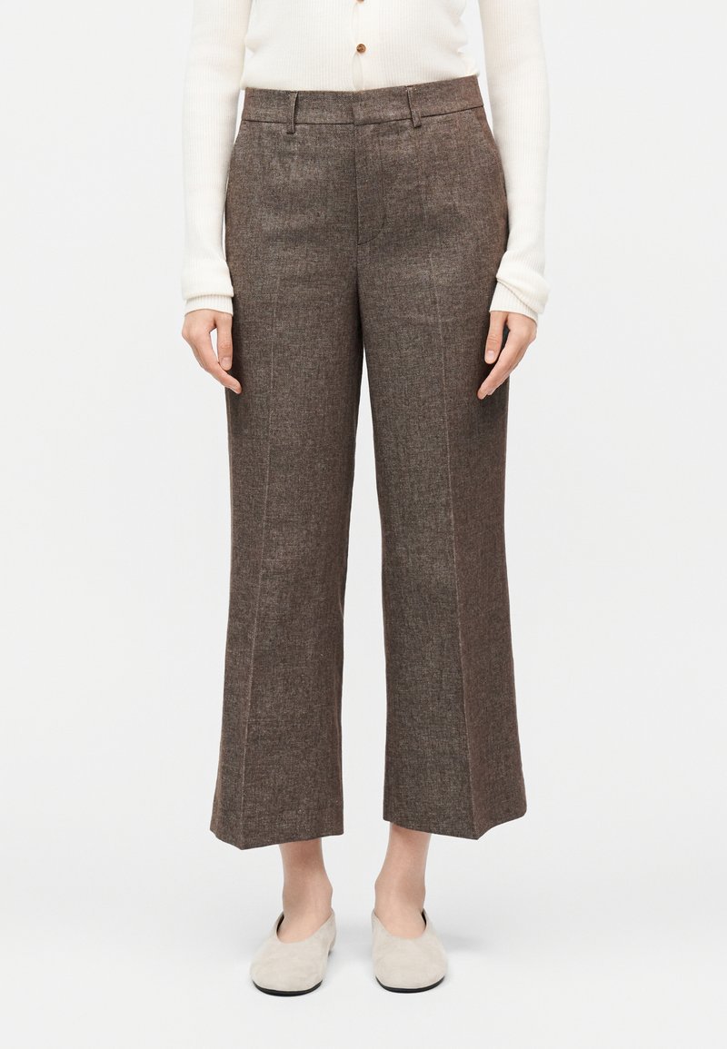 Femme portant un pantalon en tweed marron taille haute à jambes larges, un haut blanc à manches longues boutonné, et des chaussures beige slip-on, debout sur fond blanc.