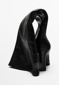 Black high-heeled shoes made of shiny leather with a flowing fabric design at the ankle. Features a sleek, curved heel and smooth texture.