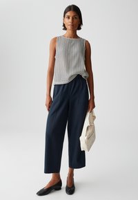 Mouwloos, gestreept topje in zwart en wit, gecombineerd met marineblauwe, wijde broekspijpen. Zwarte platte schoenen en een beige tas maken de look compleet.