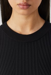 Närbild av en persons nedre ansikte och nacke som bär en svart ribbad crewneck-tröja med långt mörkt hår synligt.