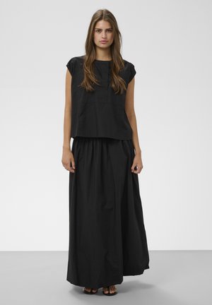 Jeune femme aux longs cheveux bruns portant un haut noir sans manches ample et une longue jupe noire, debout devant un fond uni.