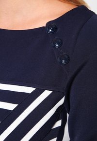 Marineblaues Oberteil mit horizontalen weißen Streifen, strukturiertem Stoff und drei glänzenden Knöpfen an der Schulter für zusätzliche Details.