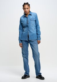 Helles blaues Denim-Hemd mit langen Ärmeln, Knopfverschluss und zwei Brusttaschen, kombiniert mit passenden Jeans und schwarzen Schuhen.