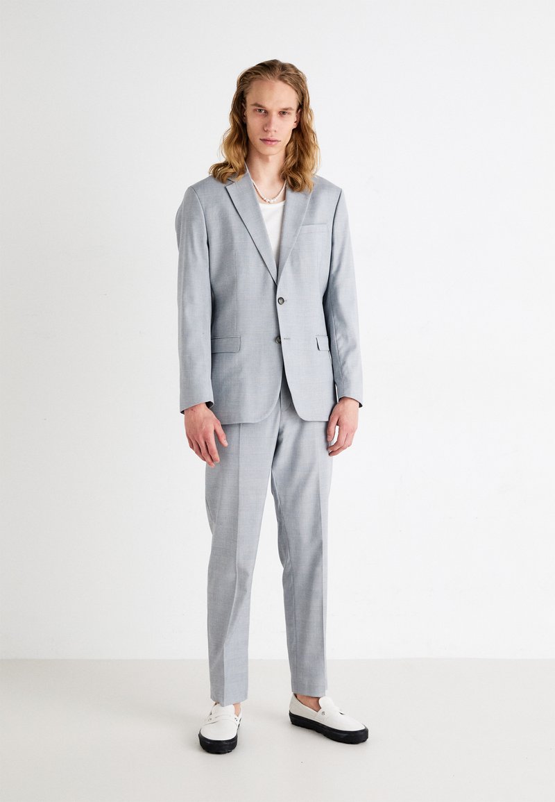 Completo grigio chiaro con blazer a un bottone e pantaloni coordinati. Indossato sopra una maglietta bianca. Abbinato a scarpe bianche con suola nera.