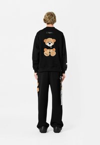 Persona con cabello rubio vistiendo sudadera negra y pantalones con diseños de ositos de peluche, de pie en posición de espaldas sobre un fondo blanco.