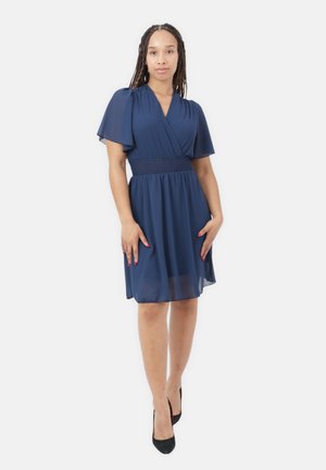 Femme aux cheveux tressés portant une robe bleu marine longueur genoux avec des manches évasées et des talons hauts noirs, debout devant un fond blanc.