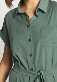 Donna che indossa un abito camicia verde con bottoni, maniche corte arrotolate, tasche sul petto e una cintura di tessuto legata in vita.