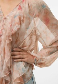 Blusa leggera e trasparente in rosa pallido con motivi floreali acquerellati, bordi arricciati e chiusure a bottoni, caratterizzata da maniche lunghe e ampie.