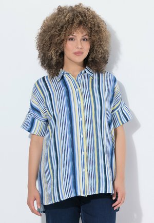 Frau mit lockigem Haar, die ein locker sitzendes kurzärmeliges Hemd mit vertikalen blauen, weißen und gelben Streifen trägt, steht an einer weißen Wand.