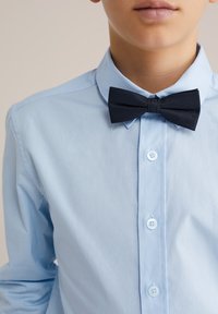 Šviesiai mėlynas marškinėlis su klasikiniu apykakliu, vertikaliu sagų užsegimu ir juodu lanku iš tekstūruotos medžiagos.