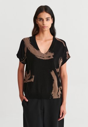 Femme aux longs cheveux foncés portant un haut noir à manches courtes en V avec des motifs abstraits marron et un pantalon noir, debout devant un fond blanc.
