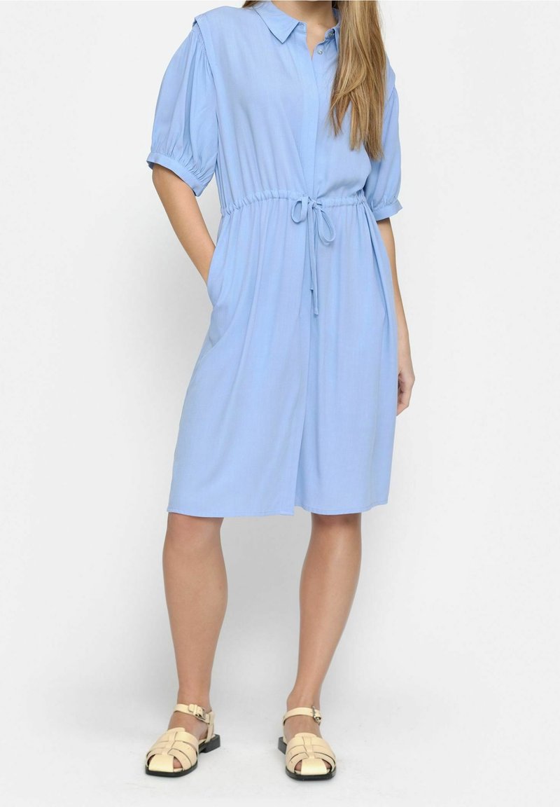 Femme portant une robe bleu clair jusqu'aux genoux avec des manches courtes bouffantes, un col, une taille ajustée avec un cordon, et des sandales beige à bout fermé.