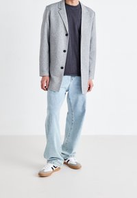 Uomo che indossa un blazer grigio chiaro, una maglietta grigio scuro, jeans azzurri chiari e sneakers bianche con strisce nere su sfondo bianco.