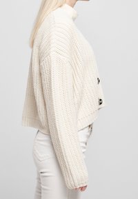 Tjock, krämfärgad stickad cardigan med en kortad design, ribbad nederkant och tre stora mörka knappar fram.