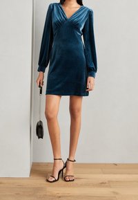 Sametové tyrkysově modré šaty s hlubokým V výstřihem, dlouhými balónovými rukávy a přiléhavým střihem. V páru s černými sandály na podpatku a malou černou kabelkou.