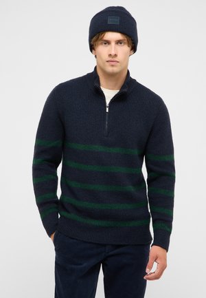Man wearing a navy blue knit sweater with green stripes and a half-zip, paired with a navy blue beanie and dark pants.
