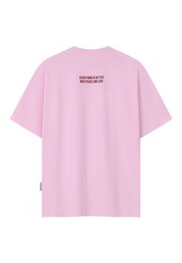 T-shirt rose clair à manches courtes avec un texte rouge sur le haut du dos indiquant « TOUT EST MIEUX AVEC LA PAIX ET L'AMOUR » et une petite étiquette latérale.