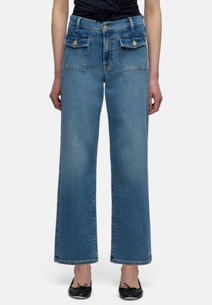 Woman wearing high-waist blue wide-leg jeans with front flap pockets and black ballet flats, standing against plain background.