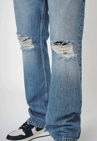 Svetle modré distressed džíny s velkými vytrhnutými otvory na kolenou a třepenými okraji, s volným střihem a bledou texturou látky.