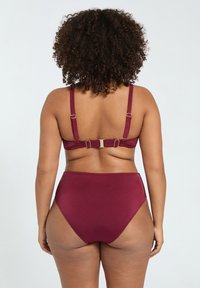 Persona con cabello rizado vista desde atrás que lleva un bikini de dos piezas color granate con tirantes ajustables y bragas de cintura alta.