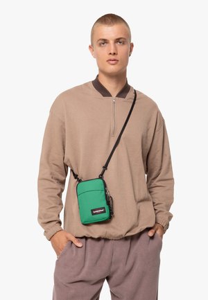 Sacoche verte avec fermeture éclair noire et branding, portée en travers du corps. Modèle en sweat beige et pantalon gris, avec un design minimaliste.