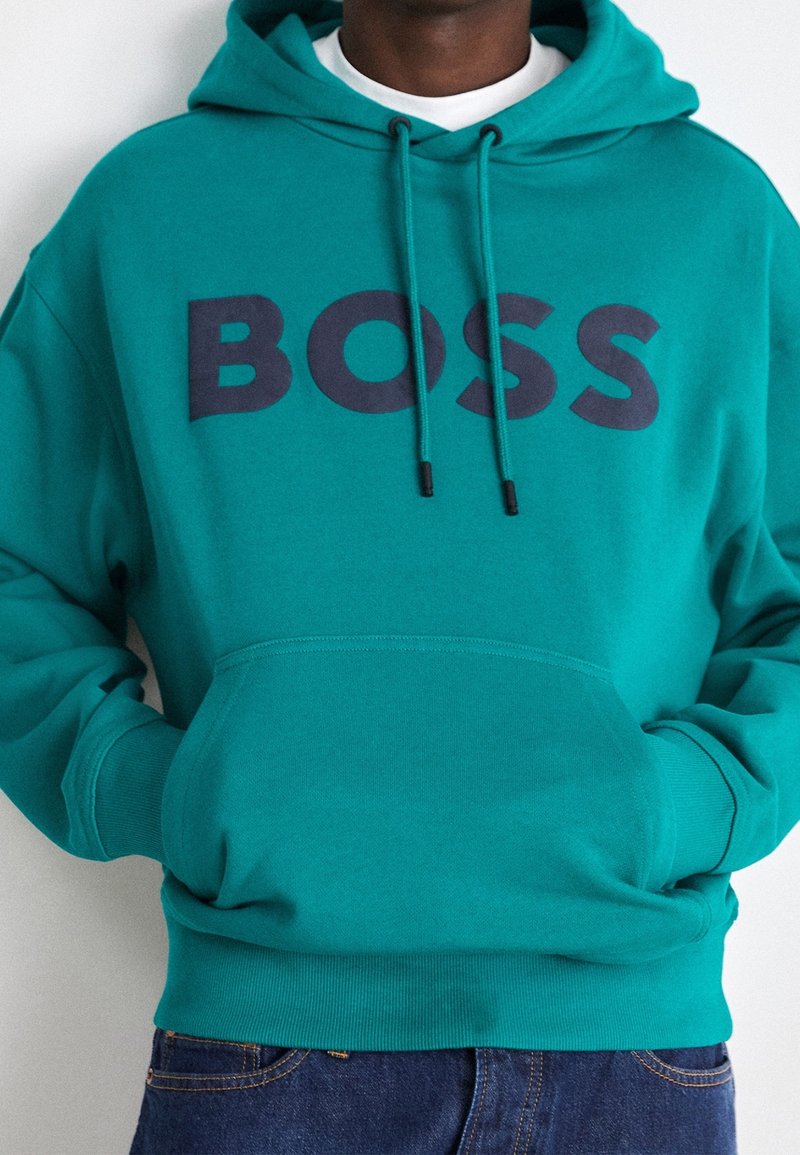 Homme portant un sweat à capuche turquoise avec poche avant et texte noir "BOSS" sur la poitrine, associé à un jean bleu foncé.
