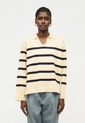 Person iført en cremet langærmet top med sorte vandrette striber og lyseblå jeans, stående vendt fremad mod en ensfarvet baggrund.