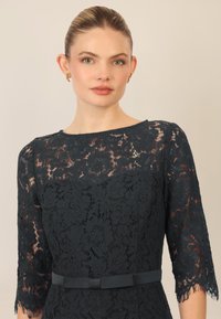 Vestido de encaje en azul marino oscuro con mangas de tres cuartos, patrón floral, escote redondo y cinturón de tela en la cintura; diseño texturizado y elegante.