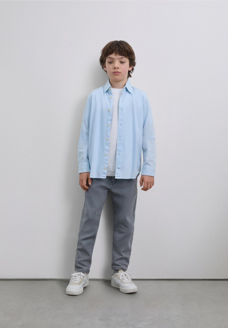 Chemise bleu clair boutonnée, T-shirt blanc en dessous, pantalon gris et baskets blanches, avec une silhouette simple et décontractée et une coupe droite.