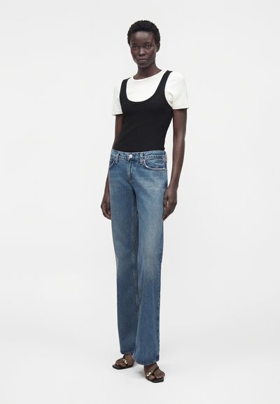 Γυναίκα στέκεται φορώντας λευκό κοντομάνικο πουκάμισο κάτω από μαύρο αμάνικο τοπ, μπλε τζιν παντελόνι με ίσιο μπατζάκι και μαύρα σανδάλια με χρυσές λεπτομέρειες.