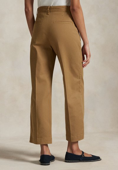 Személy, aki magas derekú, egyenes szárú tevészegszínű nadrágot és tengerészkék Mary Jane balerinát visel, semleges háttér előtt áll.