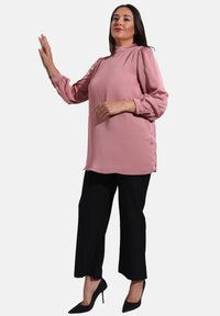 Blusa rosa a maniche lunghe con spalle arricciate, abbinata a pantaloni neri a gamba larga. La modella indossa tacchi alti neri e posa con una mano sollevata.