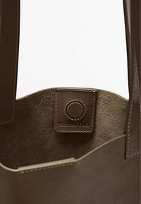 Borsa tote in suede marrone con bordi lisci, dotata di un'etichetta in pelle impressa con "Massimo Dutti" e un accento circolare.