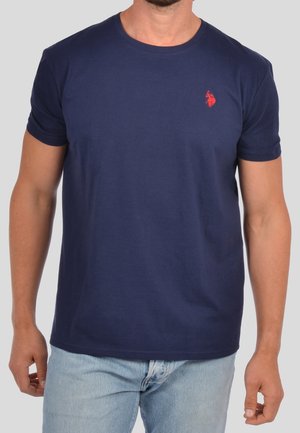 Mann trägt ein schlichtes marineblaues Kurzarm-T-Shirt mit einem kleinen roten Pferdelogo auf der linken Brust und hellblaue Jeans.