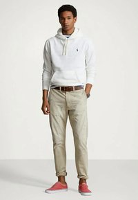 Νεαρός άνδρας που στέκεται φορώντας μια λευκή κουκούλα, μπεζ παντελόνι, καφέ ζώνη και κόκκινα αθλητικά παπούτσια, μπροστά σε απλή φωτεινή φόντο.