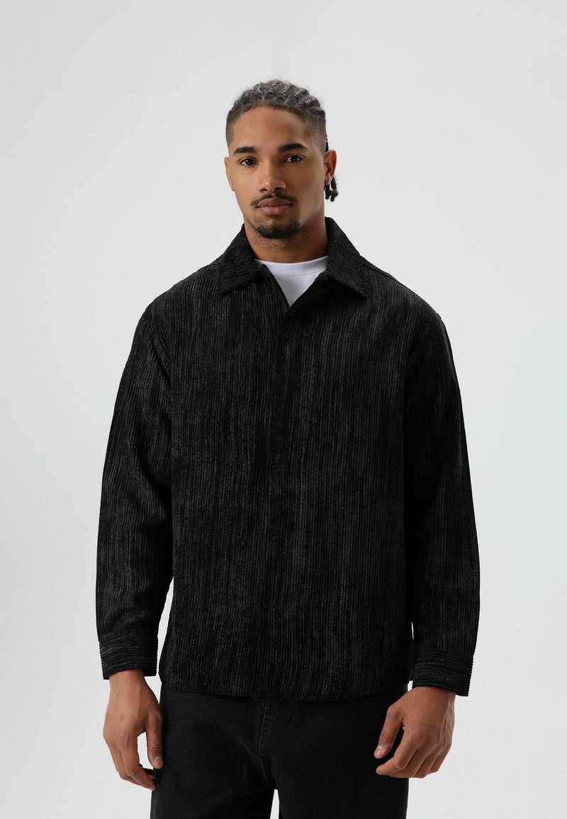 Zwart corduroy overhemd met verticale geribbelde structuur, lange mouwen, knoopsluiting aan de voorkant en kraag, gedragen over een wit T-shirt.
