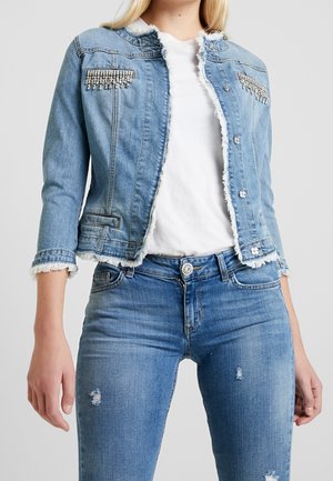 Woman wearing light blue denim jacket with fringed edges and rhinestone embellishments over a white shirt and ripped blue jeans.
