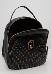 Mochila negra de imitación cuero con un patrón en chevron de tachuelas doradas. Presenta una cremallera dorada y un detalle del logotipo en el bolsillo frontal.