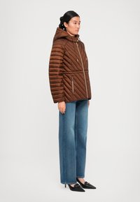 Kvinne i en brun quiltet hettejakke, vide blå jeans og svarte spisse hæler, som står foran en hvit bakgrunn.