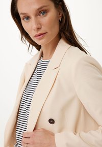 Blazer in lino pesca con revers a punta, chiusura a doppio bottone marrone, indossato sopra una camicia a righe bianco e nero. Texture liscia.