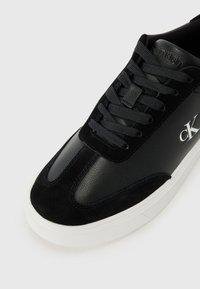Black leather and suede sneaker with flat laces, white rubber sole, and a logo detail on the side. Smooth and textured material contrast.