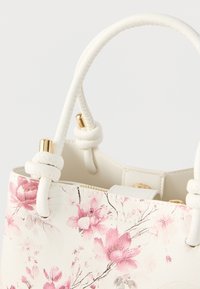 Bolso de cuero blanco con estampado floral rosa, detalles dorados y asas anudadas sobre un fondo claro.