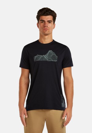 Mężczyzna ubrany w czarną koszulkę z niebieskim wzorem gór w postaci siatki drucianej oraz beżowe spodnie, stojący na białym tle.