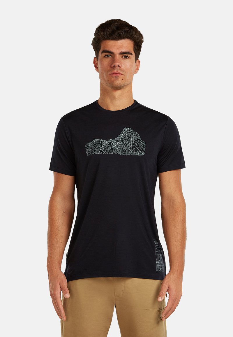 Mężczyzna ubrany w czarną koszulkę z niebieskim wzorem gór w postaci siatki drucianej oraz beżowe spodnie, stojący na białym tle.