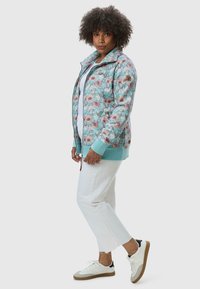 Giacca azzurro chiaro con motivo floreale, dotata di polsini e orlo elasticizzati, abbinata a pantaloni bianchi a gamba dritta e sneakers bianche con suola beige.